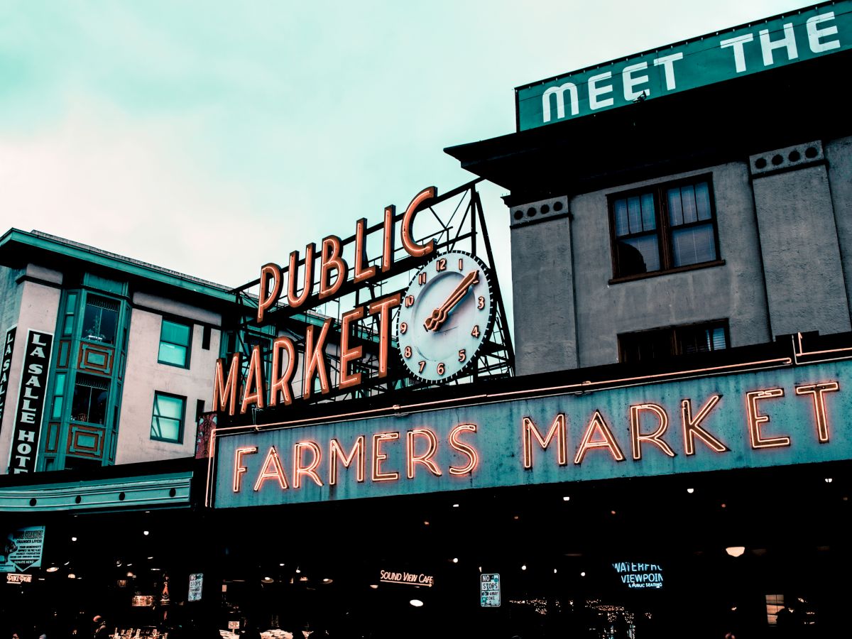 The image shows a neon sign for a "Public Market" and "Farmers Market," set against buildings with a vintage, urban atmosphere.