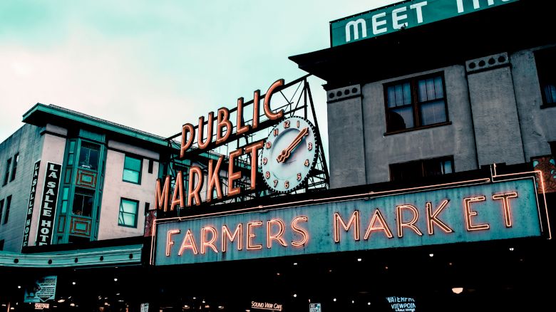 The image shows a neon sign for a "Public Market" and "Farmers Market," set against buildings with a vintage, urban atmosphere.