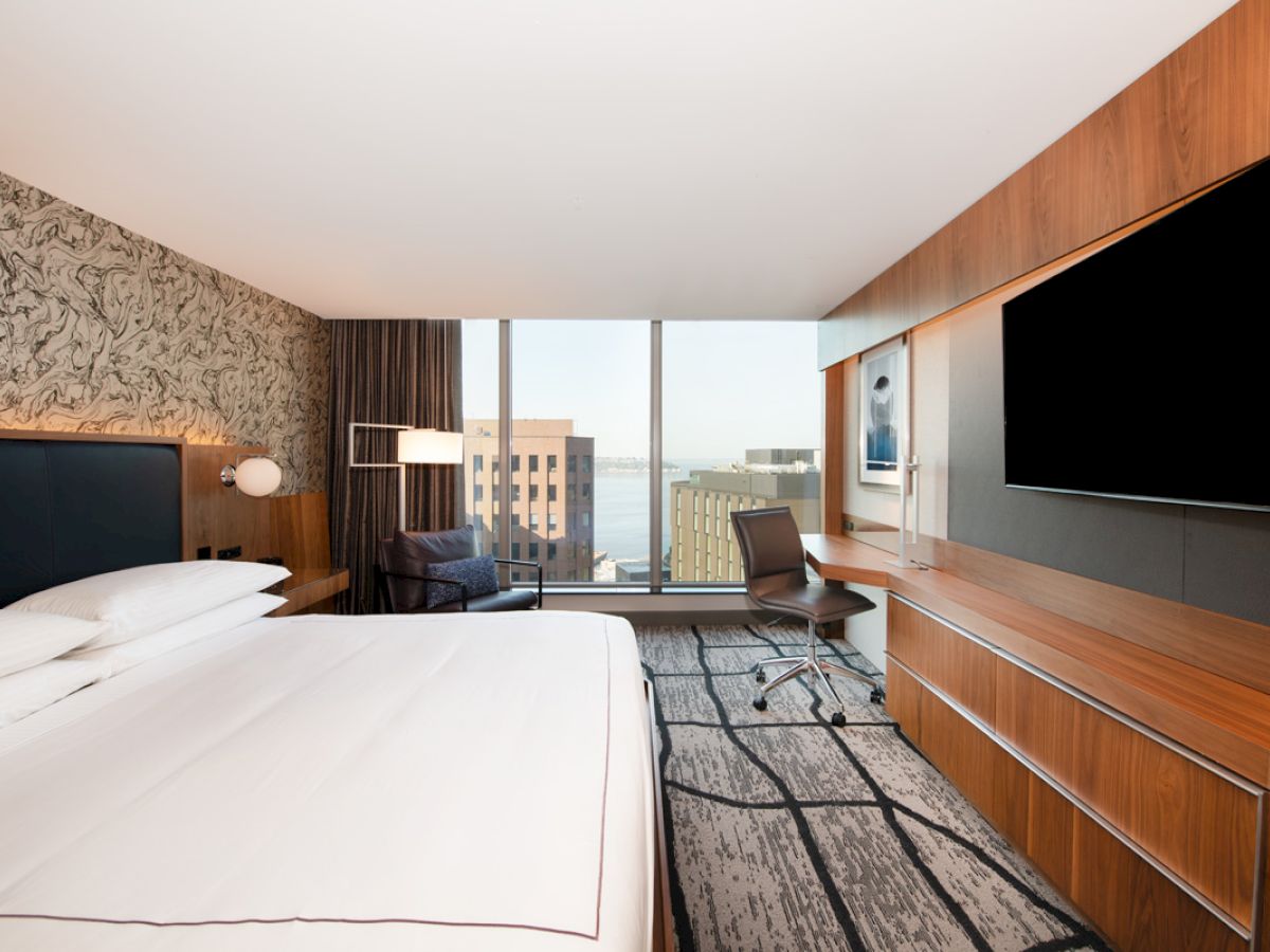 A modern hotel room with a large bed, wall-mounted TV, desk, chair, and a window view of buildings and water.