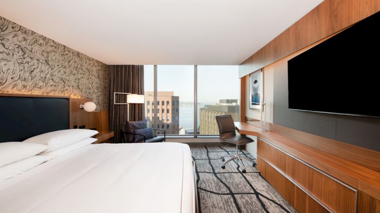 A modern hotel room with a large bed, wall-mounted TV, desk, chair, and a window view of buildings and water.