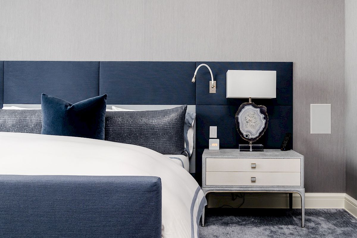 A modern bedroom with a blue upholstered bed, gray and white bedding, a lamp, and a small nightstand on a gray carpet.