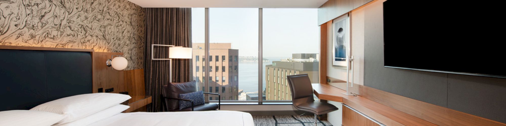 A modern hotel room with a bed, large TV, desk, and a window view of buildings and water. The decor is contemporary with wood accents.